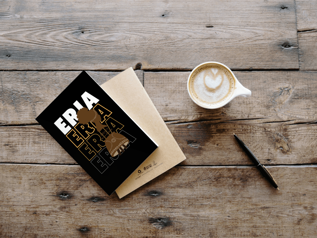 A notebook with a black cover featuring the text 'ERIA' in bold and yellow is placed on a wooden table alongside a latte with a heart-shaped design in the foam, a brown envelope, and a black pen.